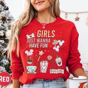 A woman wearing a red sweater with a cartoon-style design and text printed on it, standing in front of a Christmas tree.