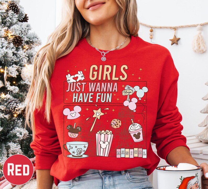 A woman wearing a red sweater with a cartoon-style design and text printed on it, standing in front of a Christmas tree.