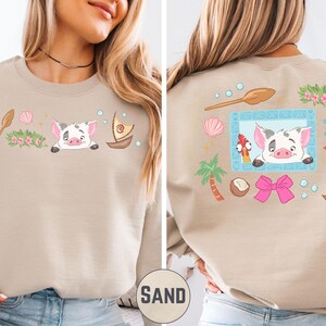 a woman wearing a beige sweater with a colorful and whimsical design on the back. The design includes a pig, a boat, a palm tree, and various other elements, creating a playful and cheerful appearance.