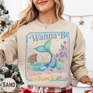 A woman wearing a beige sweater with a graphic design featuring a mermaid, castle, and text that says "I Wanna Be Where The People Aren't".