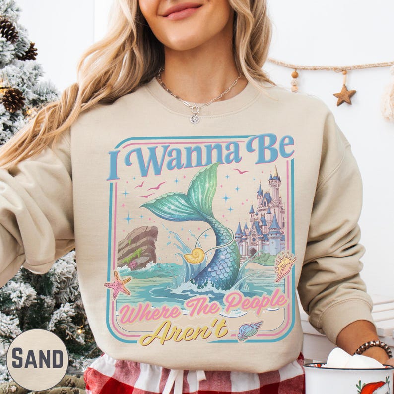 A woman wearing a beige sweater with a graphic design featuring a mermaid, castle, and text that says "I Wanna Be Where The People Aren't".