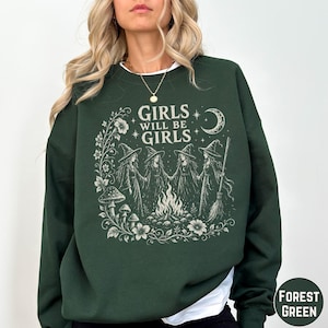 May include: A forest green crewneck sweatshirt featuring a cream-colored graphic print of four witches in pointed hats standing around a campfire. The design includes floral elements, mushrooms, a crescent moon, and the text Girls Will Be Girls.