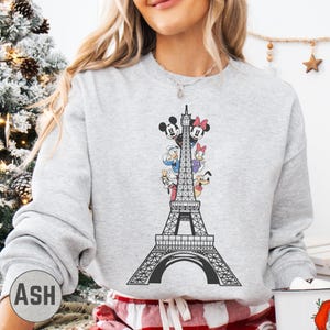 A woman wearing a gray sweatshirt with a graphic of the Eiffel Tower and Disney characters on it.