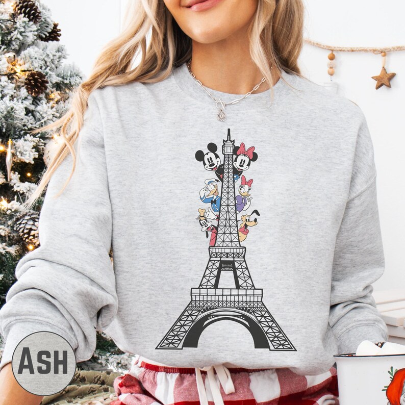 A woman wearing a gray sweatshirt with a graphic of the Eiffel Tower and Disney characters on it.