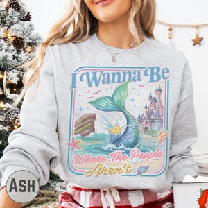 A woman wearing a gray sweatshirt with a colorful graphic design featuring a mermaid, castle, and text.