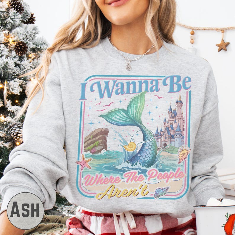 A woman wearing a gray sweatshirt with a colorful graphic design featuring a mermaid, castle, and text.