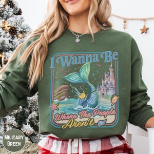 A woman wearing a green sweatshirt with a graphic design featuring a mermaid and a castle.