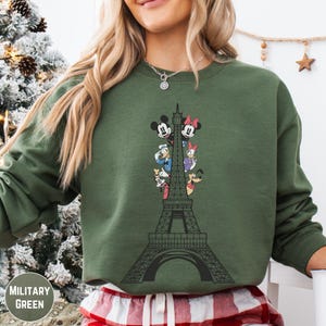 A woman wearing a green sweatshirt with a graphic of the Eiffel Tower and Disney characters on it.