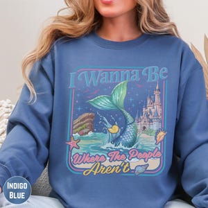 A woman wearing a blue sweatshirt with a graphic design featuring a mermaid, a castle, and the text "I Wanna Be Where The People Aren't".