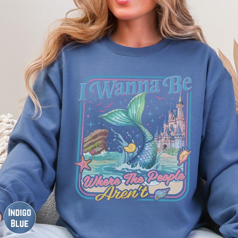 A woman wearing a blue sweatshirt with a graphic design featuring a mermaid, a castle, and the text "I Wanna Be Where The People Aren't".