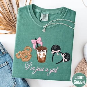 a green t-shirt with a graphic design of a cup with a pink ribbon, a pair of sunglasses, and a pretzel. The text on the t-shirt says "I'm just a girl".