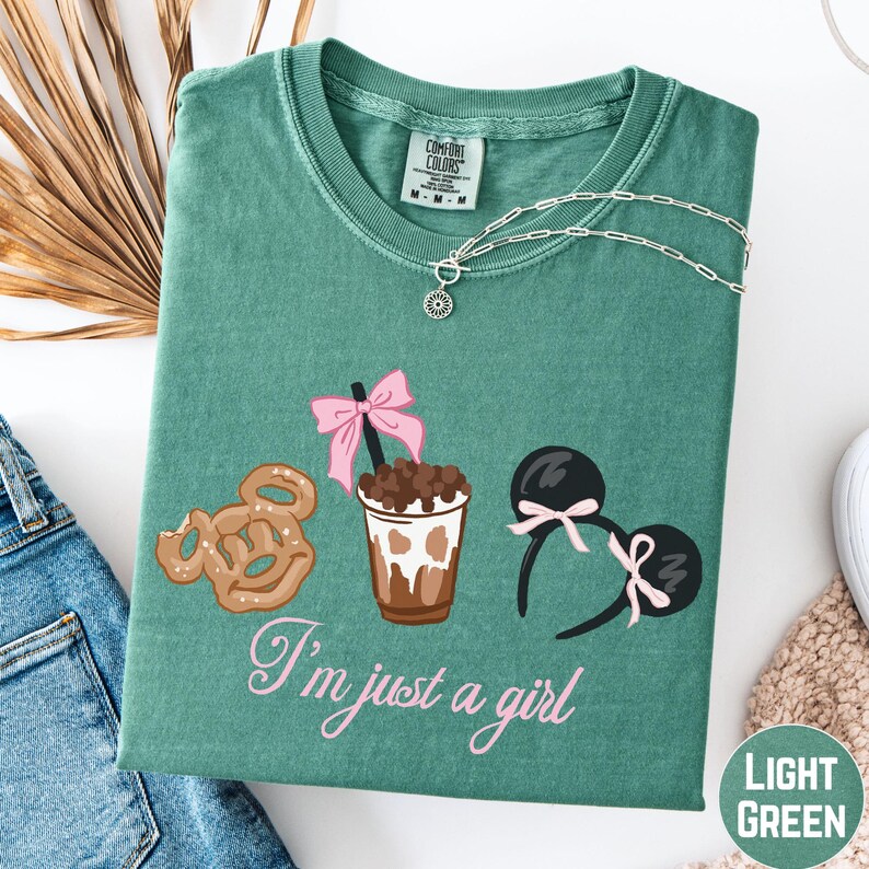 a green t-shirt with a graphic design of a cup with a pink ribbon, a pair of sunglasses, and a pretzel. The text on the t-shirt says "I'm just a girl".