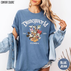 A woman wearing a blue t-shirt with a Disney World logo and text, along with denim jeans and a denim jacket.