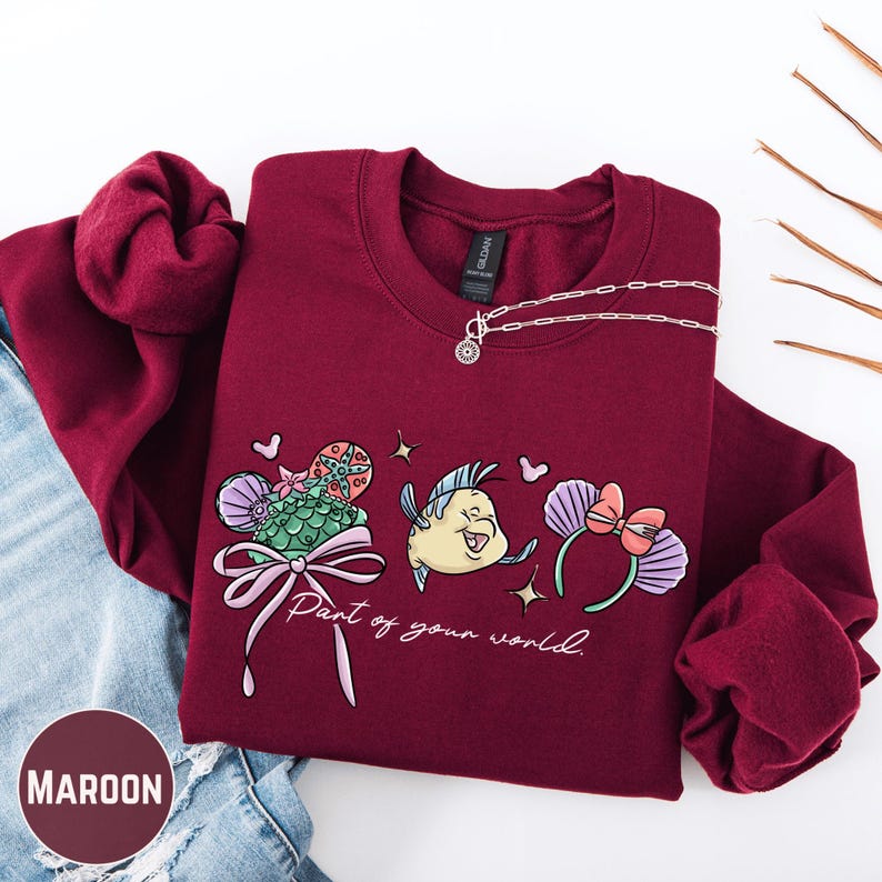 A maroon sweatshirt with a graphic design featuring a mermaid and the text "Part of your world" is displayed on a white background.