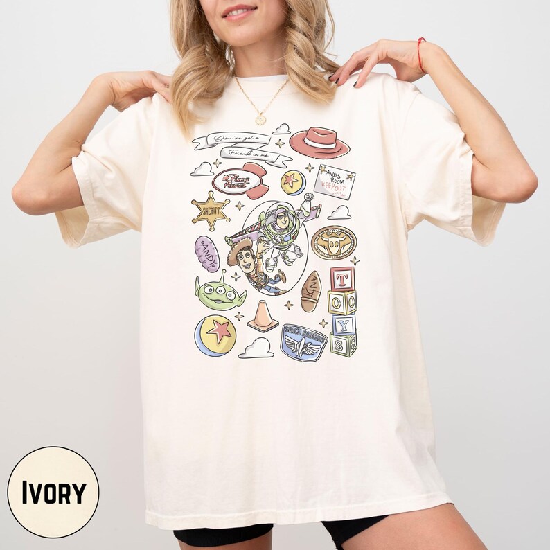 A woman wearing a white t-shirt with various cartoon and toy-related icons and text printed on it.