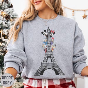 A woman wearing a gray sweatshirt with a graphic of the Eiffel Tower and Disney characters on it.