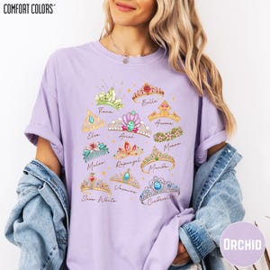 A woman wearing a purple t-shirt with a graphic design of various crowns and tiaras, along with the names of Disney princesses. She is also wearing a denim jacket and has her hair styled in loose waves.