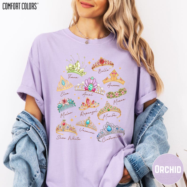 A woman wearing a purple t-shirt with a graphic design of various crowns and tiaras, along with the names of Disney princesses. She is also wearing a denim jacket and has her hair styled in loose waves.