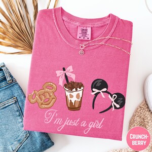 a pink t-shirt with a graphic design of a cup with a drink, a pair of Mickey Mouse ears, and a donut. The text on the t-shirt says "I'm just a girl".