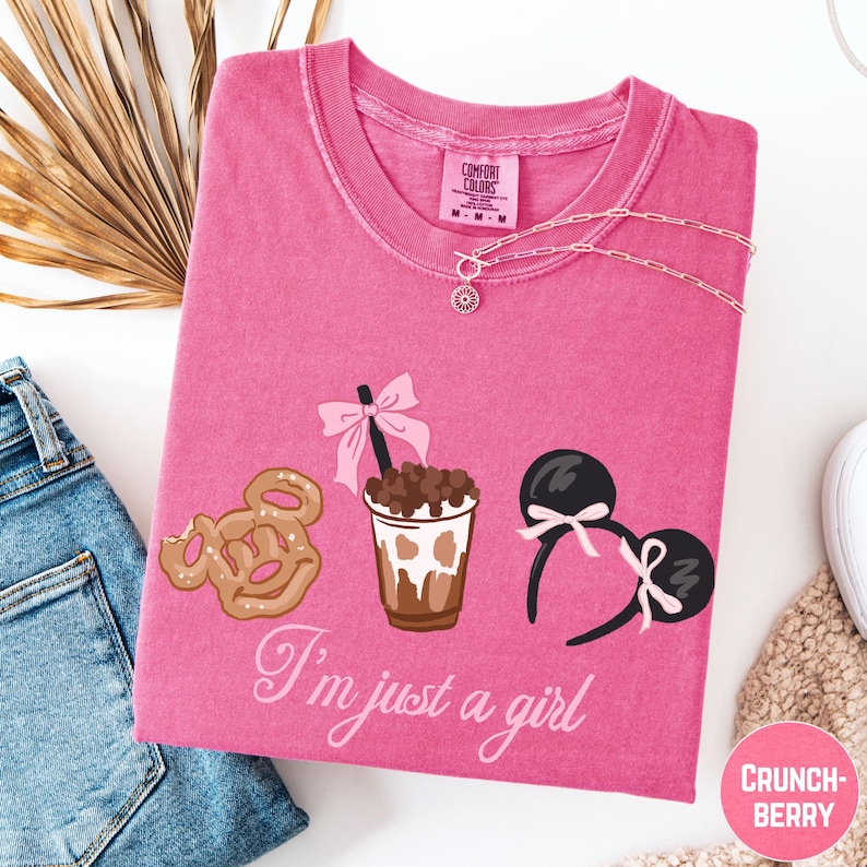 a pink t-shirt with a graphic design of a cup with a drink, a pair of Mickey Mouse ears, and a donut. The text on the t-shirt says "I'm just a girl".