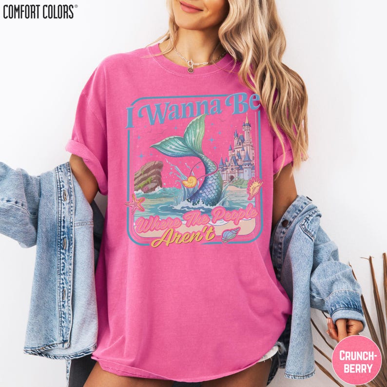 A woman wearing a pink t-shirt with a mermaid graphic and text, along with a denim jacket, stands against a white background.