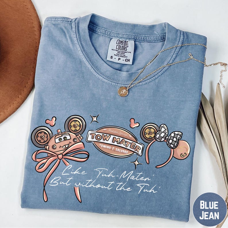 A blue t-shirt with a cartoon character design and text, along with a necklace and a brown hat, placed on a white surface.