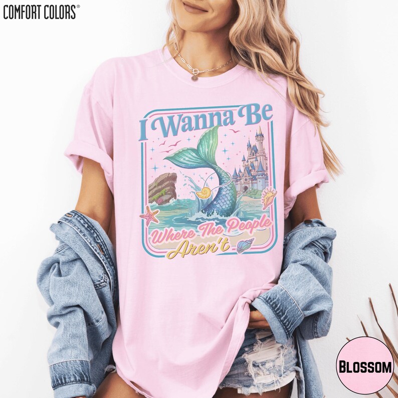 A woman wearing a pink t-shirt with a mermaid graphic and text, along with a denim jacket, posing against a white background.