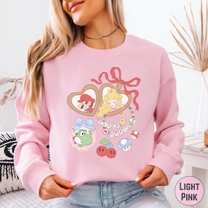 May include: Light pink sweatshirt featuring a cartoon design with Mario and Princess Peach in heart-shaped lockets. The design includes Yoshi, cherries, and mushrooms. The text "PEACH MARIO" is also included in the design.