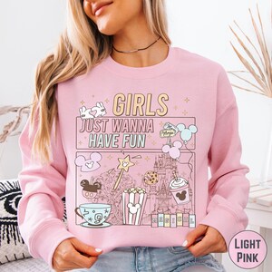 A woman wearing a pink sweatshirt with a graphic design featuring various Disney-themed illustrations, including Mickey Mouse, popcorn, and a castle.