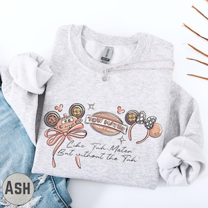 A gray sweatshirt with a graphic design featuring cartoon-style characters and text, along with a necklace and a pair of jeans.