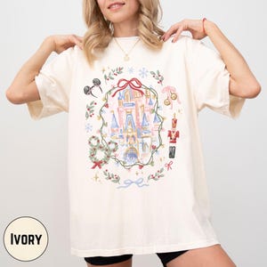 May include: Ivory-colored t-shirt featuring a whimsical castle design within a wreath of greenery, bows, and snowflakes. The design includes Mickey Mouse ears, a toy soldier, and ornaments. The word "IVORY" is printed at the bottom.