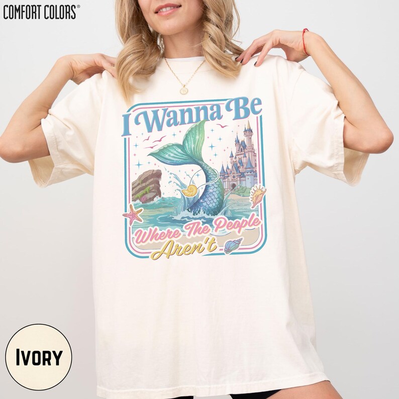 A woman wearing a white t-shirt with a colorful graphic design featuring a mermaid and a castle.