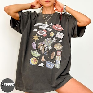 A person wearing a black t-shirt with a graphic design featuring various cartoon characters and icons, including a character from the Toy Story franchise.