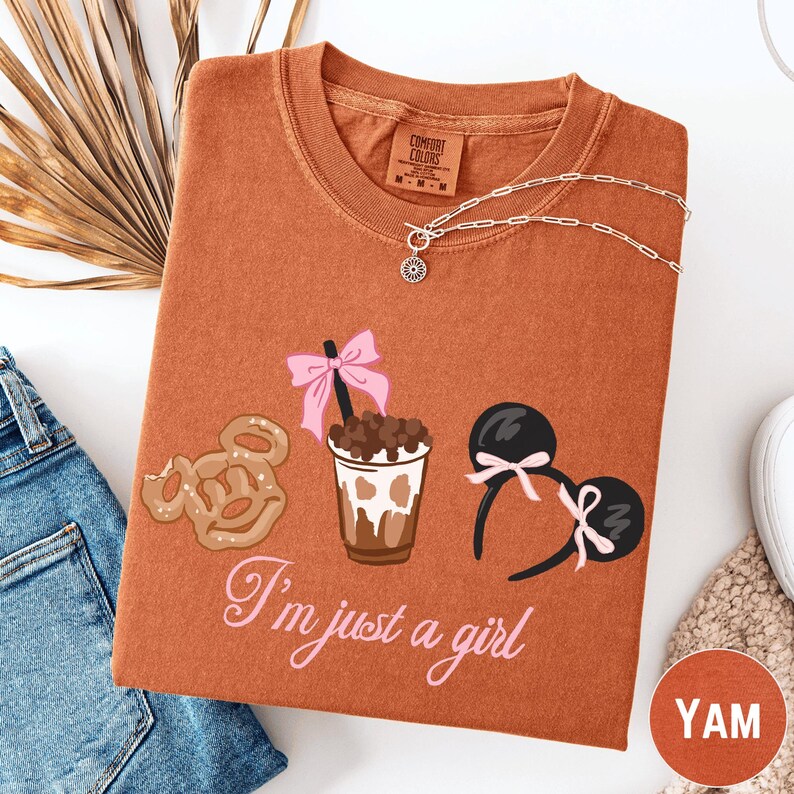 a brown t-shirt with a graphic design of a cup with a beverage, a pair of Mickey Mouse ears, and a donut. The text on the t-shirt says "I'm just a girl".