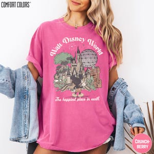 A woman wearing a pink t-shirt with a graphic of Disney World and the text "The happiest place in earth" is posing for a photo.