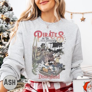 A woman wearing a gray sweatshirt with a graphic of Disney characters and the text "Pirates of the Caribbean: The Deadman's Cove" printed on it, standing in front of a Christmas tree.