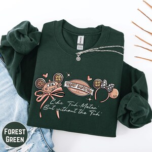 A green sweatshirt with a graphic design featuring cartoon-like characters and text, along with a necklace and a pair of jeans.