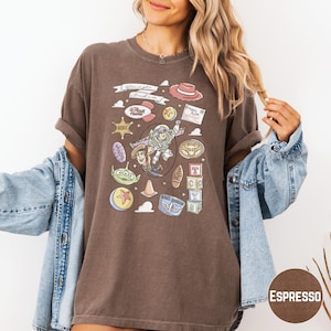 A woman wearing a brown t-shirt with various cartoon characters and icons, including a character from the Toy Story franchise, is posing with her arms outstretched.