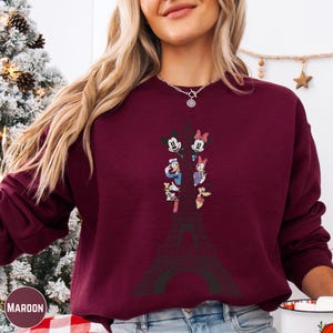 A woman wearing a burgundy sweatshirt with a graphic of the Eiffel Tower and Disney characters on it.