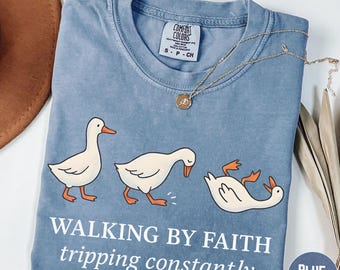 May include: Blue jean colored t-shirt with a graphic of three white cartoon geese, one tripping. The text reads "WALKING BY FAITH tripping constantly". A gold necklace is draped across the shirt.
