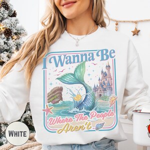 A woman wearing a white sweatshirt with a colorful graphic design featuring a mermaid, a castle, and the text "I Wanna Be Where The People Aren't".