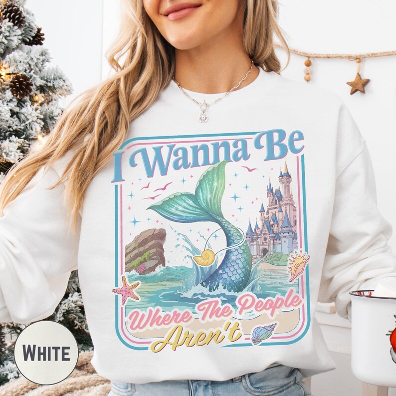 A woman wearing a white sweatshirt with a colorful graphic design featuring a mermaid, a castle, and the text "I Wanna Be Where The People Aren't".