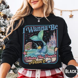 A woman wearing a black sweatshirt with a colorful graphic design featuring a mermaid, castle, and text.