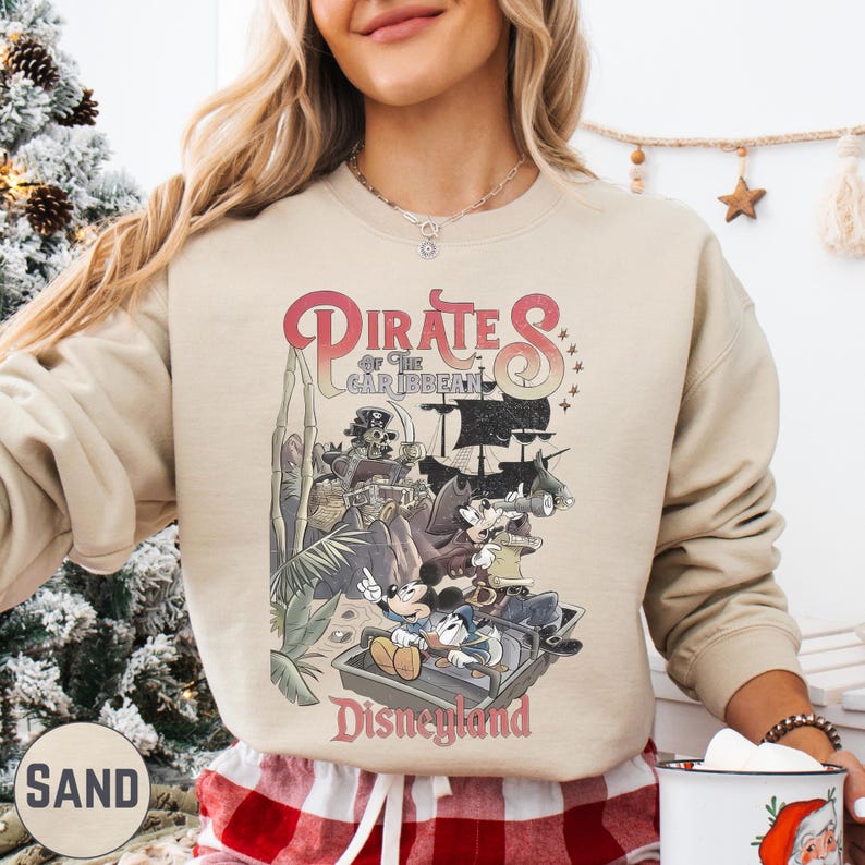 A woman wearing a beige sweatshirt with a graphic of Disney's Pirates of the Caribbean on it, standing in front of a Christmas tree.