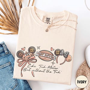a beige t-shirt with a cartoon-style design, a necklace, and some text and graphics.