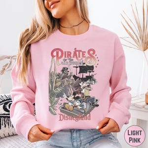 A woman wearing a pink sweatshirt with a graphic of Disney characters from the Pirates of the Caribbean ride.