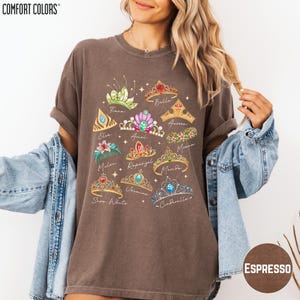 A woman wearing a brown t-shirt with a graphic design of various crowns and tiaras, along with the text "Comfort Colors" printed on it. She is also wearing a denim jacket and has her arms outstretched.