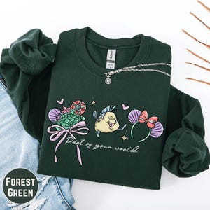 A green sweatshirt with a graphic design featuring a cartoon fish and flowers, along with the text "Part of your world" printed on it.