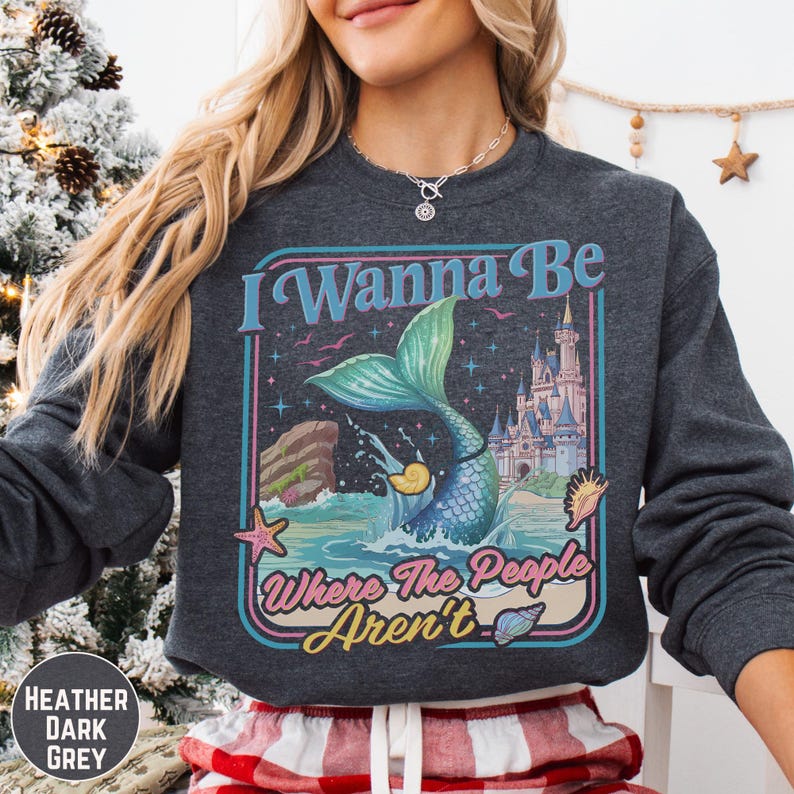 A woman wearing a gray sweatshirt with a colorful graphic design featuring a mermaid, castle, and text.