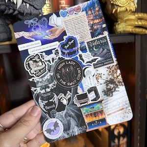 May include: A rectangular sticker sheet featuring various stickers with fantasy themes. Stickers include text like "Bat Boys", "High Lady of the Smut Court", and "Velaris". The sheet is held in a hand, with a bookshelf in the background.